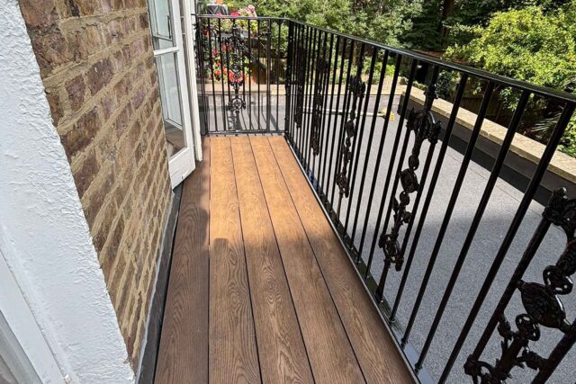 Highly engineered Millboard composite decking providing a realistic oak finish alongside structural black metal railings on a narrow London balcony.
