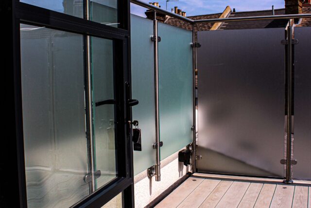 Premium composite decking laid upon a weather-proofed warm roof system, leading towards a balcony door and enclosed by a frosted glass balustrade on an urban London terrace.